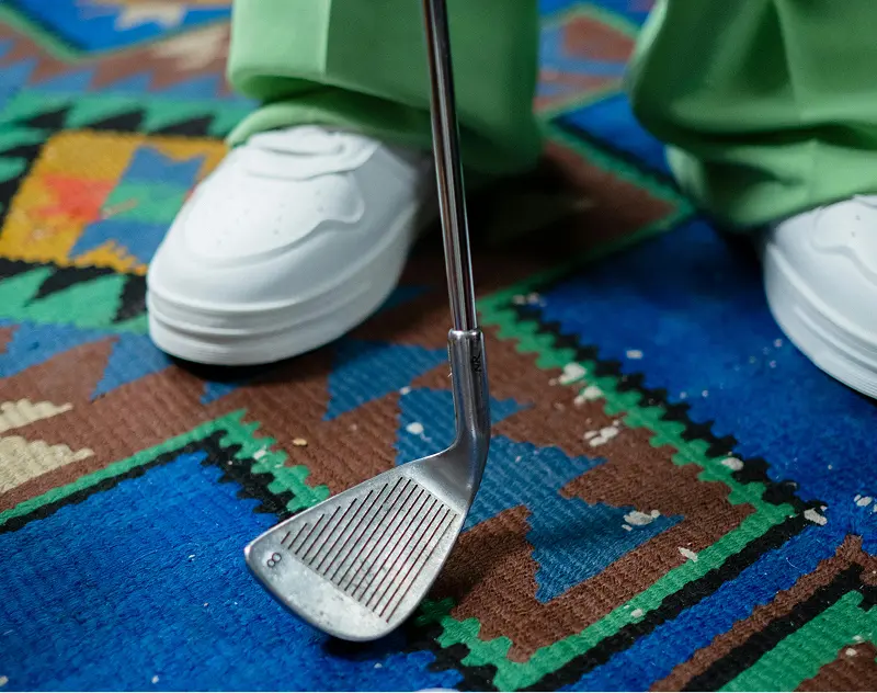 Golf club resting on a patterned rug beside a person wearing white shoes