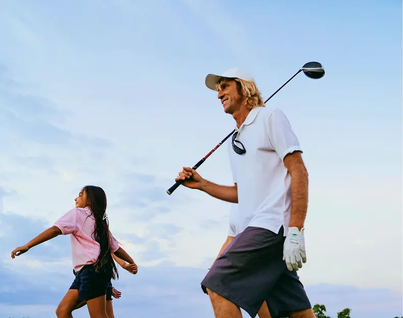 Golfer carrying a driver while walking the course with a child at sunset
